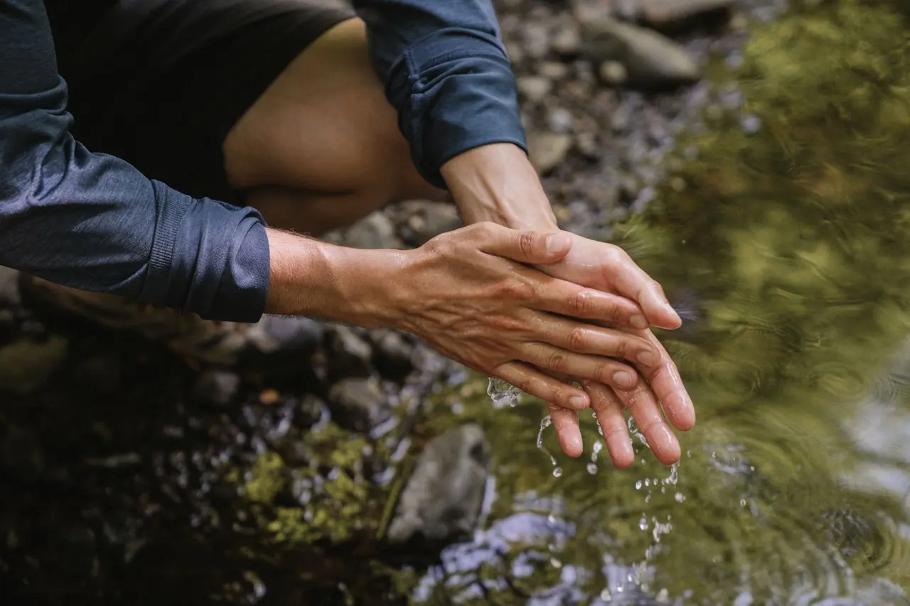 Close-up of a person crouched by a clear stream rinsing their hands, water dripping from their fingers