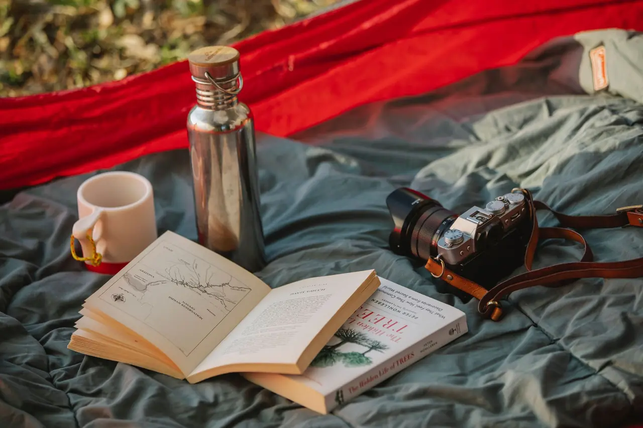 Camping gear laid out on a sleeping bag: stainless steel bottle, enamel mug, field guide books and a film camera