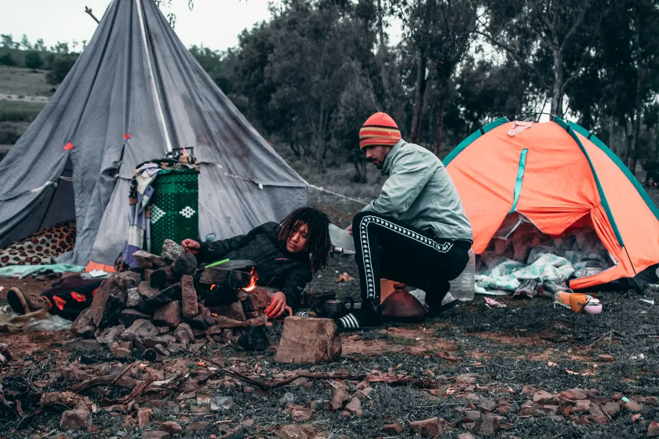Two friends tending a small campfire between a gray tipi tent and an orange dome tent at a wooded campsite
