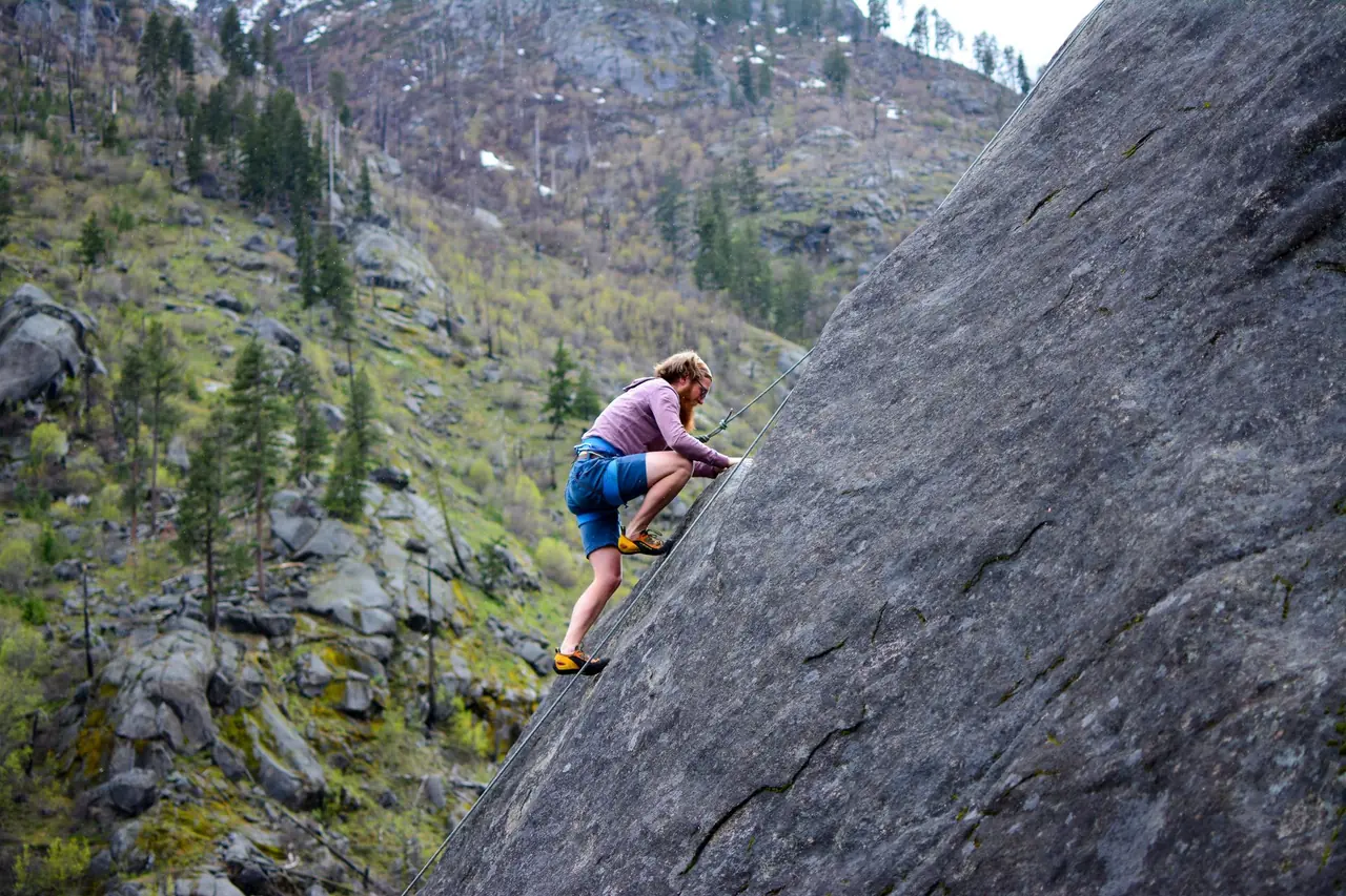Person using hands and feet to scramble up a steep granite slab on a forested mountainside