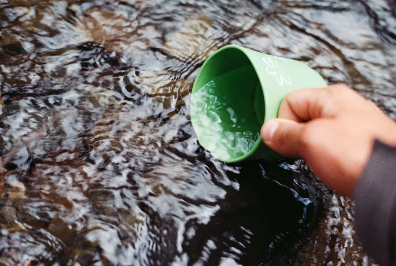Hand dipping a green camp cup into a clear, rippling stream to collect water