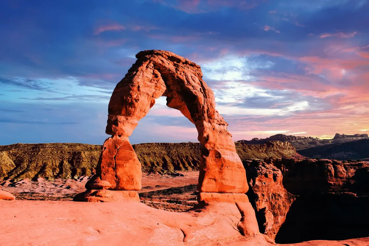 Delicate Arch standing on red slickrock under a colorful sunset sky in Arches National Park, Utah