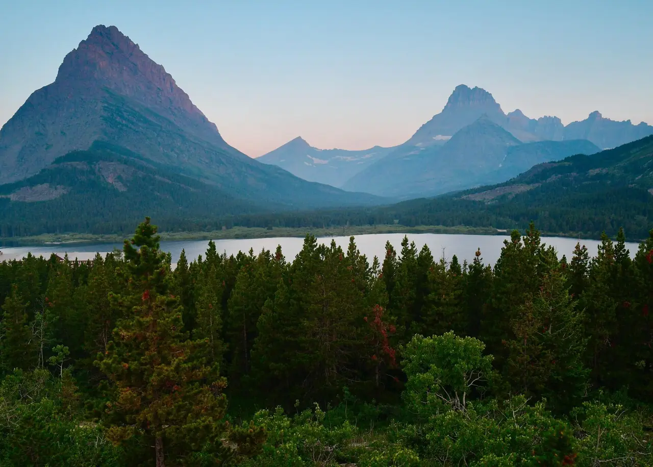 Pointed peaks of the Many Glacier area rising above a lake and dense evergreen forest at dusk in Glacier National Park, Montana