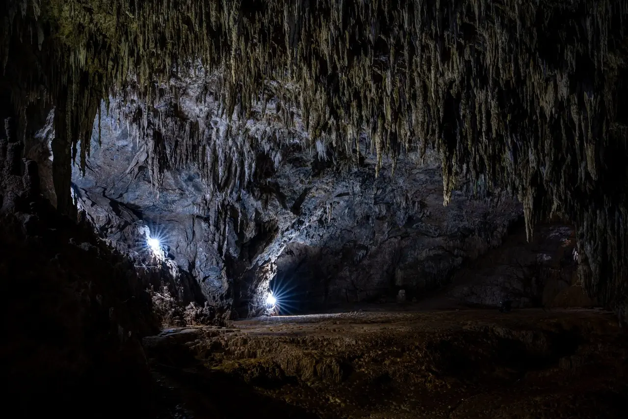 A large limestone cave chamber with a ceiling of hanging formations, lit by two small lights
